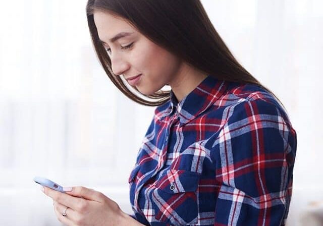 Vertical of sociable young girl texting on smartphone in living room