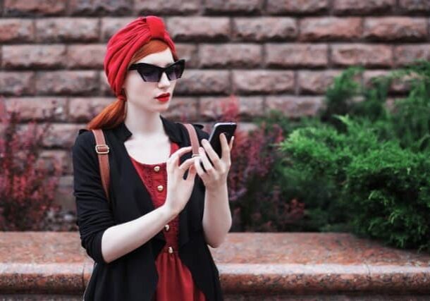 Stylish red-haired girl in sunglasses and in a red turban with a mobile phone in her hands. A woman with a new phone. Black phone in hand. The girl is looking at the phone. Internet on the phone. Touchscreen phone